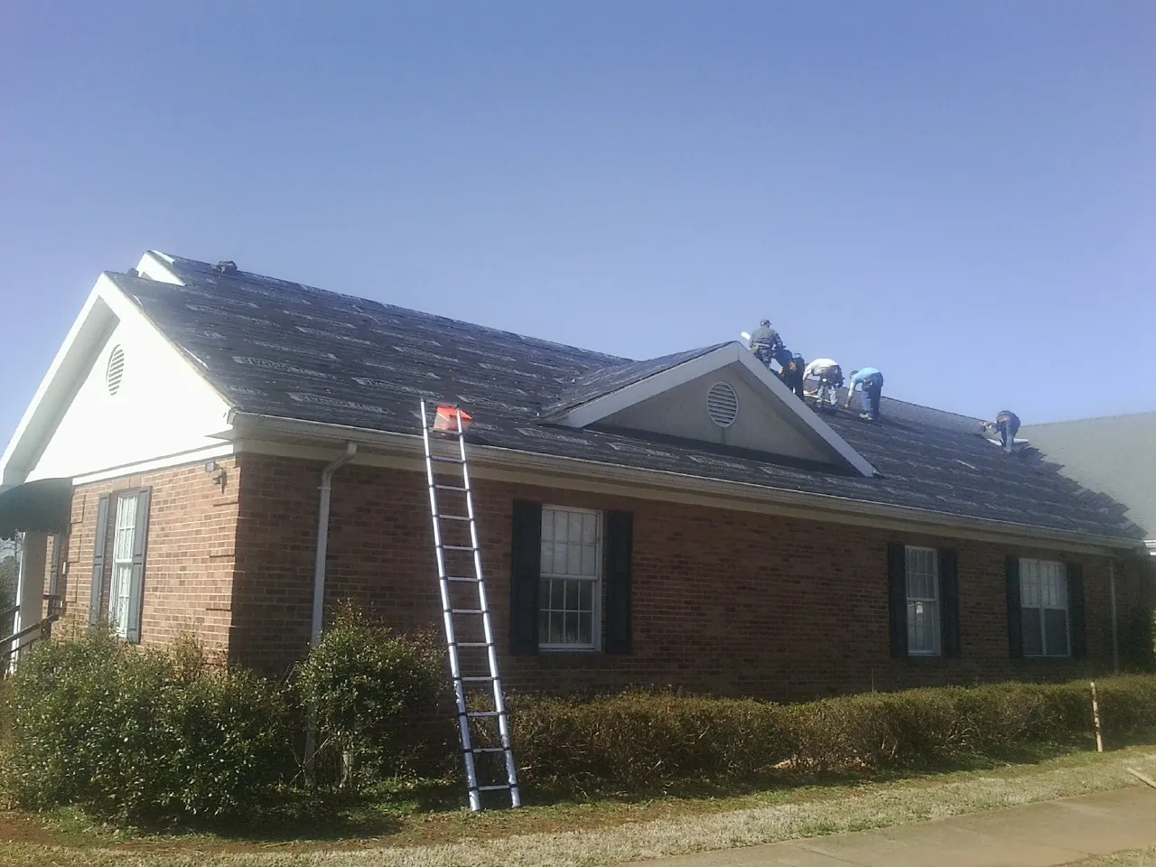 Crew tearing shingles off a brick ranch roof with a ladder set — Metal Roof Repair work by our crew in Sleepy Eye