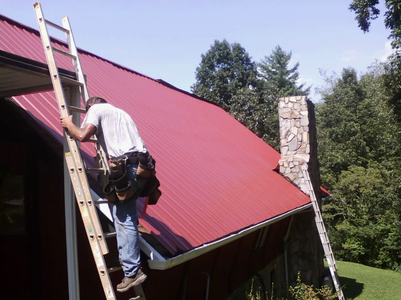 Roofer on a ladder beside a red metal roof and stone chimney — Soffit & Fascia Repair by Roofing Brothers in Sleepy Eye